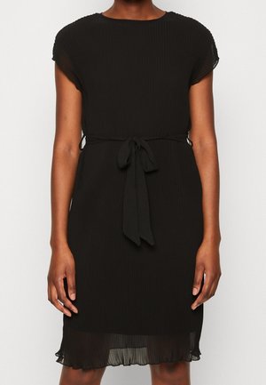 Femme portant une robe plissée noire longueur genou avec des manches courtes et une ceinture en tissu nouée à la taille.