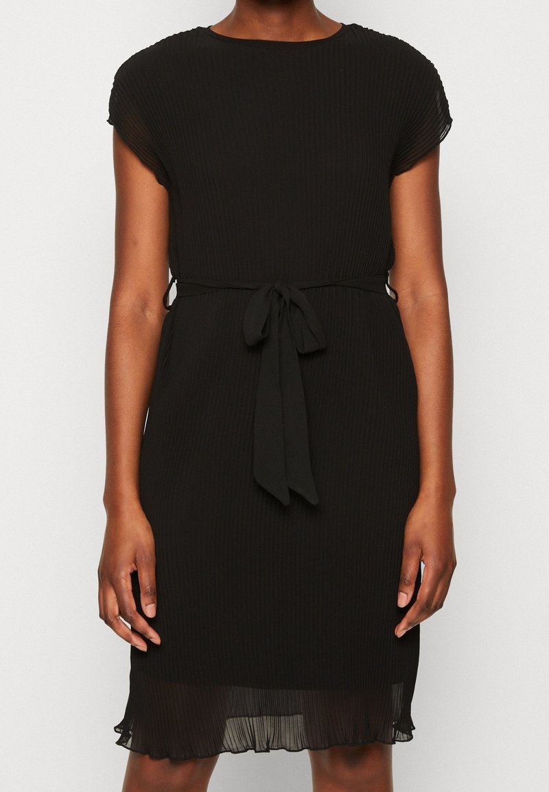Femme portant une robe plissée noire longueur genou avec des manches courtes et une ceinture en tissu nouée à la taille.