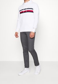 Bílá mikina s kapucí s černými a červenými vodorovnými pruhy, logo na hrudi; v kombinaci se šedými úzkými kalhotami a bílými teniskami.