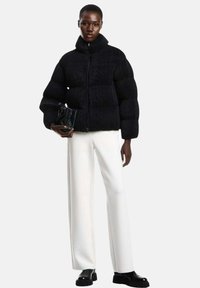 Giacca a vento nera con superficie testurizzata, colletto alto e chiusura con zip; abbinata a pantaloni bianchi a gamba larga e scarpe nere. Tiene una borsa piccola.