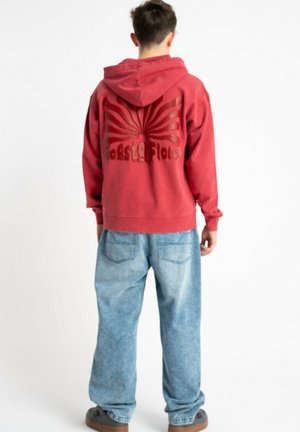 Osoba w czerwonej bluzie z kapturem z promienistym wzorem i napisem na plecach, w połączeniu z luźnymi jasnoniebieskimi dżinsami i brązowymi butami.