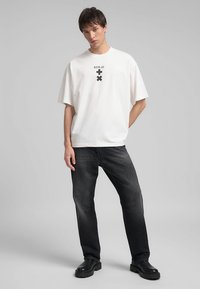T-shirt bianco oversize con logo nero stampato e croci, abbinato a jeans in denim scuro e calzature chunky nere. Sfondo semplice.