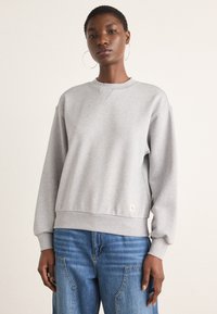 Felpa grigia con scollo a girocollo, vestibilità comoda, polsini e orlo a costine, e un discreto logo sul davanti. Abbinata a jeans blu.
