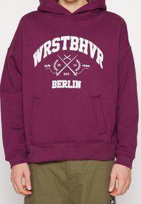 Paarse hoodie met witte grafische tekst "WRSTBHVR" en "BERLIN." Heeft een kangoeroezak en ribgebreide mouwen. Van een katoenmix.