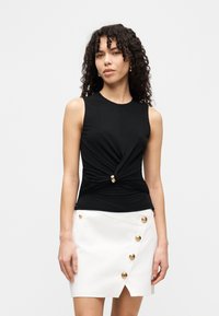 Μαύρο μπλουζάκι χωρίς μανίκια με σχεδιασμό στριφογυριστός και χρυσή λεπτομέρεια στη μέση, σε συνδυασμό με μια λευκή φούστα περιτυλίγματος με χρυσά κουμπιά.