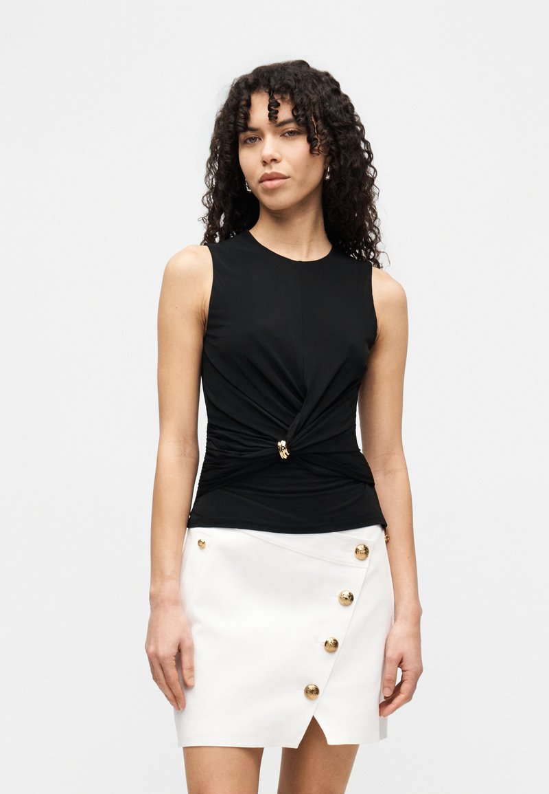 Μαύρο μπλουζάκι χωρίς μανίκια με σχεδιασμό στριφογυριστός και χρυσή λεπτομέρεια στη μέση, σε συνδυασμό με μια λευκή φούστα περιτυλίγματος με χρυσά κουμπιά.