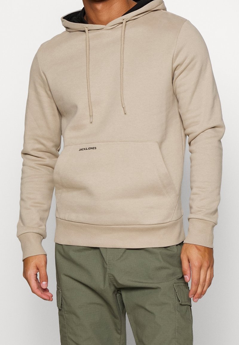 Sudadera beige con cordones, bolsillo frontal, puños acanalados y dobladillo; presenta el logotipo "JACK&JONES" impreso cerca del bolsillo.
