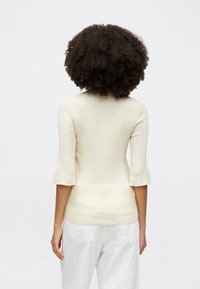 Top en tricot beige clair avec des manches évasées trois quarts, affichant une silhouette ajustée et une texture douce. Porté avec un pantalon blanc ample.
