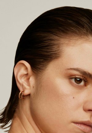 Pendiente de aro de oro con un diseño entrelazado que presenta pequeños acentos elevados. Colocado sobre un tono de piel suave y claro y cabello oscuro.