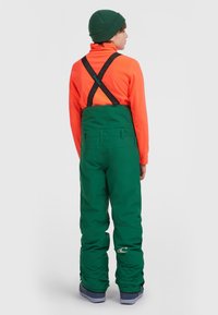Pantalones de nieve verdes con tirantes negros ajustables, combinados con una parte superior de color naranja brillante. Presenta un corte holgado, textura suave y detalle de logotipo.