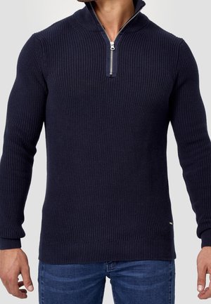 Man draagt een marineblauwe geribbelde gebreide trui met een halve ritssluiting bij de kraag en een blauwe spijkerbroek, staand tegen een effen achtergrond.