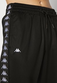 Pantalon de survêtement noir avec taille élastique et cordon de serrage, arborant des logos blancs Kappa le long du côté et un sur la cuisse avant.