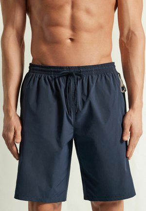 Homme torse nu portant un short de bain bleu marine avec cordon et une poche latérale zippée, debout devant un fond clair uni.