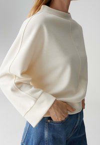 Crèmekleurige trui met een hoge halslijn, ingezakte schouders en geribbelde manchetten, gecombineerd met blauwe spijkerbroeken. Zachte stoftextuur zichtbaar.