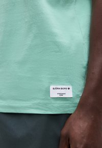 Κοντινό πλάνο του τελειώματος ενός πουκαμίσου σε πράσινο μέντας με μια λευκή ετικέτα Björn Borg και ένα σκουρόχρωμο χέρι να ακουμπά δίπλα του.