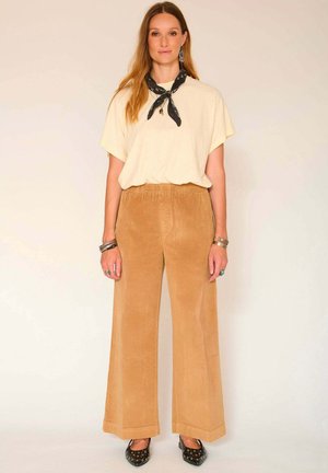 T-shirt beige oversize avec des manches retroussées, associé à un pantalon large en velours côtelé marron clair taille haute et des ballerines noires à motifs.