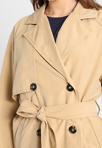 Trench-coat beige avec un design croisé, boutons noirs, ceinture à nouer à la taille, et larges revers en tissu doux et lisse.