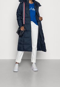 Tmavě modrý prošívaný kabát s uzávěrem na zip a druky, s kapucí; ve spojení s modrým tričkem, bílými kalhotami a bílými teniskami. Černá kabelka přes rameno.