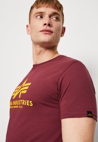Bordo kokvilnas t-krekls ar dzeltenu grafisku logo priekšā, īsām piedurknēm un mazu logo birku uz piedurknes. Gluda tekstūra.