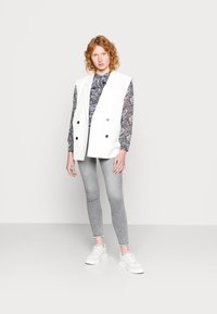 Colete branco de dois botões sobre uma camisa estampada, combinado com jeans skinny cinza e ténis brancos. Tecido suave, corte descontraído.