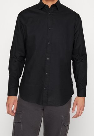 Man draagt een zwart overhemd met lange mouwen en knopen en donkergrijze cargobroek, staand tegen een effen achtergrond.