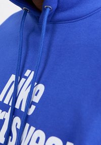 Blå hoodie med vit text. Innehåller justerbar huva och mjuk textur, med Nike-logotypen tydligt framhävd på framsidan.