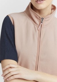 Fleeceväst i mjuk rosa med hög krage och kort ärm i marinblått ribbat tyg. Har en framficka med dragkedja och en tygdragning.