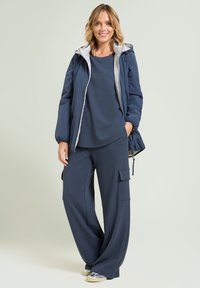 Mujer de pie, vestida con una chaqueta con capucha azul marino sobre una blusa a juego y pantalones anchos con bolsillos de carga, y zapatillas plateadas.
