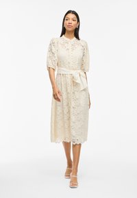 Spetsklänning i blek krämfärg, med en figurnära bodice, korta puffärmar, knäppning framtill och ett knytband i midjan för ytterligare definition.