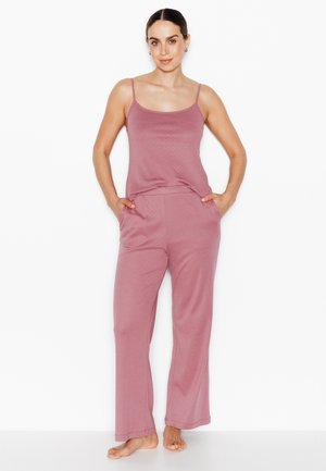 Femme aux cheveux foncés, chignon bas, portant un haut sans manches mauve et un pantalon large assorti, debout pieds nus, mains dans les poches.