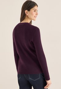 Gerippter Strickpullover in tiefem Violett mit langen Ärmeln und Rundhalsausschnitt. Gestylt mit blauer Denim-Hose, die eine strukturierte Oberfläche und eine figurbetonte Form zeigt.