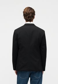 Άνδρας με κοντά σγουρά καστανά μαλλιά που φοράει μαύρο σακάκι και μπλε τζιν, ορατός από πίσω σε λευκό απλό φόντο.