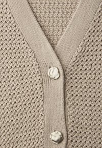 Beige gebreid vest met een getextureerd patroon en twee grote onregelmatige metalen knopen op de voorpaskant.