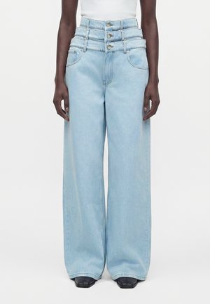 Person iført højtaljede, lyseblå, vidde jeans med tre synlige knapper, sat sammen med sorte sko med firkantet tå.