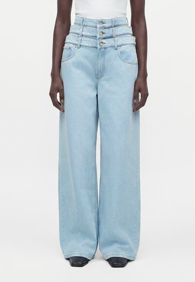 Personne portant un jean taille haute bleu clair, à jambes larges, avec trois boutons visibles, associé à des chaussures noires à bout carré.