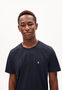 T-shirt bleu marine en coton, avec un col rond et des manches courtes. Comprend un petit logo en ruban blanc sur le côté gauche de la poitrine.