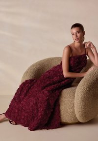 Bordeauxkleurige bloemenjurk met gelaagde rozenapplicaties, mouwloos ontwerp en een vrij vallende pasvorm die over een gestructureerde, beige gebogen stoel hangt.