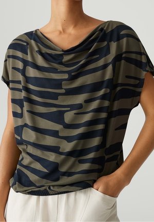 Mujer con una camiseta holgada de manga corta con rayas abstractas en azul marino y verde oliva, combinada con pantalones blancos, mano en el bolsillo.