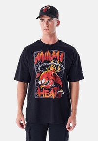 T-shirt nera oversize con grafica di una fiamma rossa e gialla rappresentante un pesce, accompagnata dalla scritta "MIAMI HEAT". Abbinata a un cappellino nero con logo.