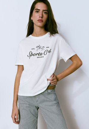 Camiseta de algodón blanca con un estampado gráfico que dice "Club Deportivo" con un diseño de caballo. Mangas cortas y corte relajado. Pantalones a rayas visibles.