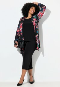 Zwarte bloemen kimono met lange mouwen over een zwarte top en cropped broek. Plat zwart hakken en een zwarte handtas. Soepele stof met een levendig bloemenpatroon.