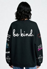 Čierna mikina s voľným strihom. Na prednej strane nápis "buď láskavý" bielou farbou. Rukávy sú zdobené farebnými grafickými prvkami a textom, vrátane srdiečok a vzorov.
