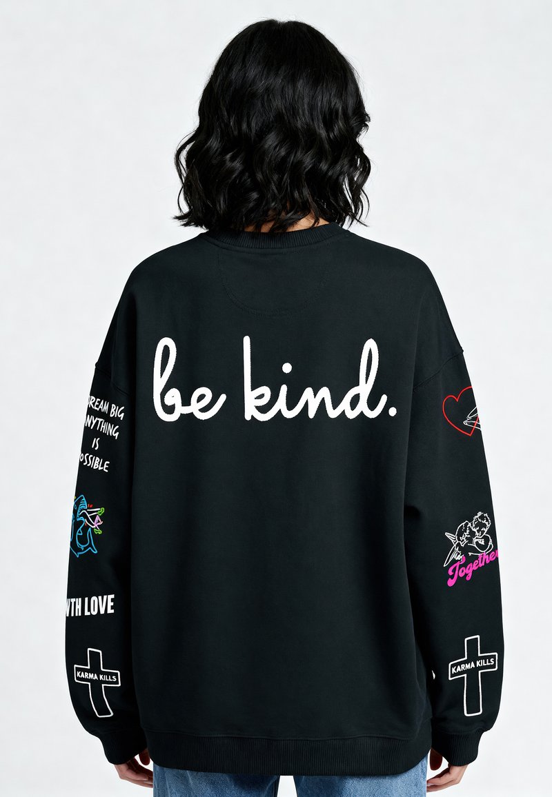 Čierna mikina s voľným strihom. Na prednej strane nápis "buď láskavý" bielou farbou. Rukávy sú zdobené farebnými grafickými prvkami a textom, vrátane srdiečok a vzorov.