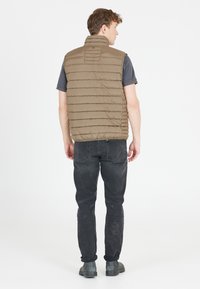 Bruine gewatteerde vest met een hoge kraag, achternaaddetails, gedragen over een korteblodig donkergrijs shirt, gecombineerd met zwarte jeans en grijze laarzen.