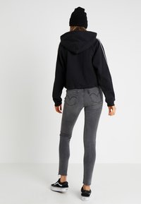 Felpa nera tagliata con cappuccio e strisce bianche sui lati, abbinata a jeans skinny grigi a vita alta e sneakers nere.