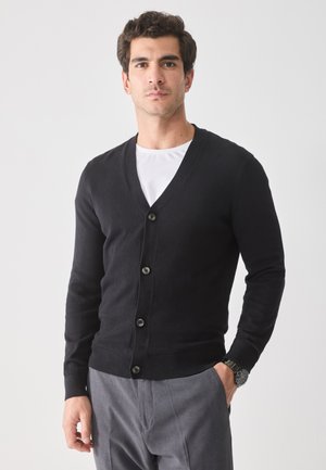Black cardigan with a V-neck, long sleeves, and five buttons. Knitted material, ribbed cuffs and hem. Worn over a white shirt.