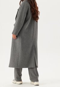 Manteau long gris à coupe droite, fabriqué en matière texturée, avec une fente au dos, accompagné d'un pantalon gris et de baskets.