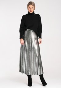 Czarny sweter z golfem w połączeniu ze srebrną, metaliczną, plisowaną suknią maxi z dopasowaną talią i miękką teksturą. Look dopełniają czarne botki sięgające kostek.