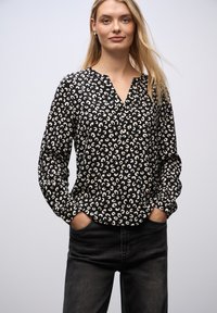 Sort bluse med V-hals, lange ærmer og løs pasform, prydet med et kremfarvet abstrakt leopardprint. Modellen har mørke jeans på.