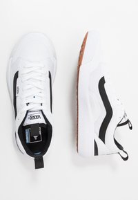 Weiße Sportschuhe mit schwarzen seitlichen Akzenten, strukturiertem Mesh-Obermaterial und Gummisohle mit einem gemusterten Profil. Zunge mit Logo sichtbar.